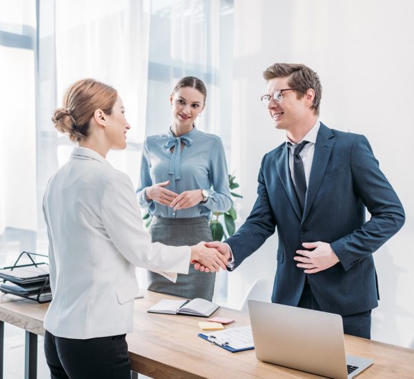 cheerful-recruiter-shaking-hands-with-woman-near-colleague-in-office