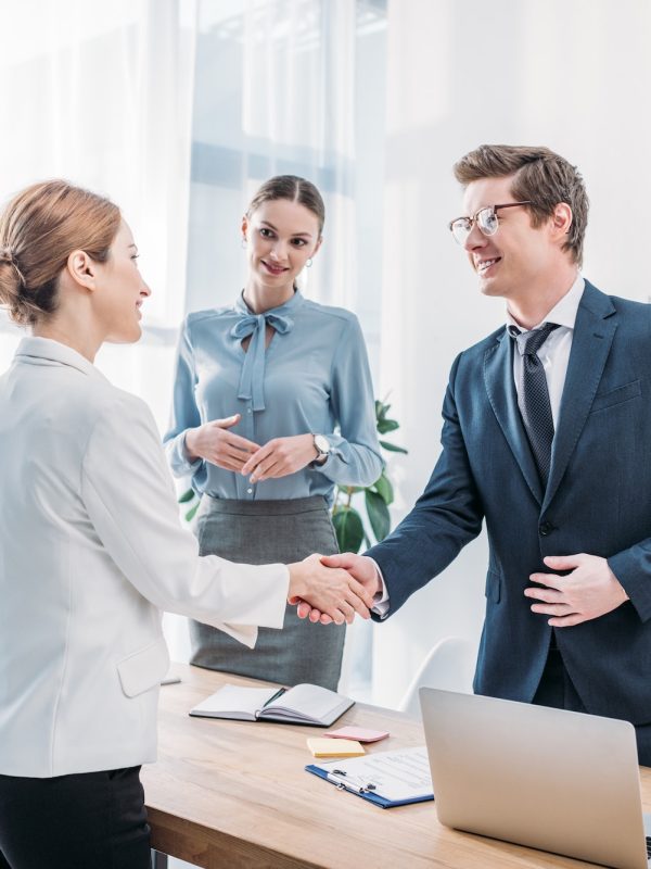 cheerful-recruiter-shaking-hands-with-woman-near-colleague-in-office