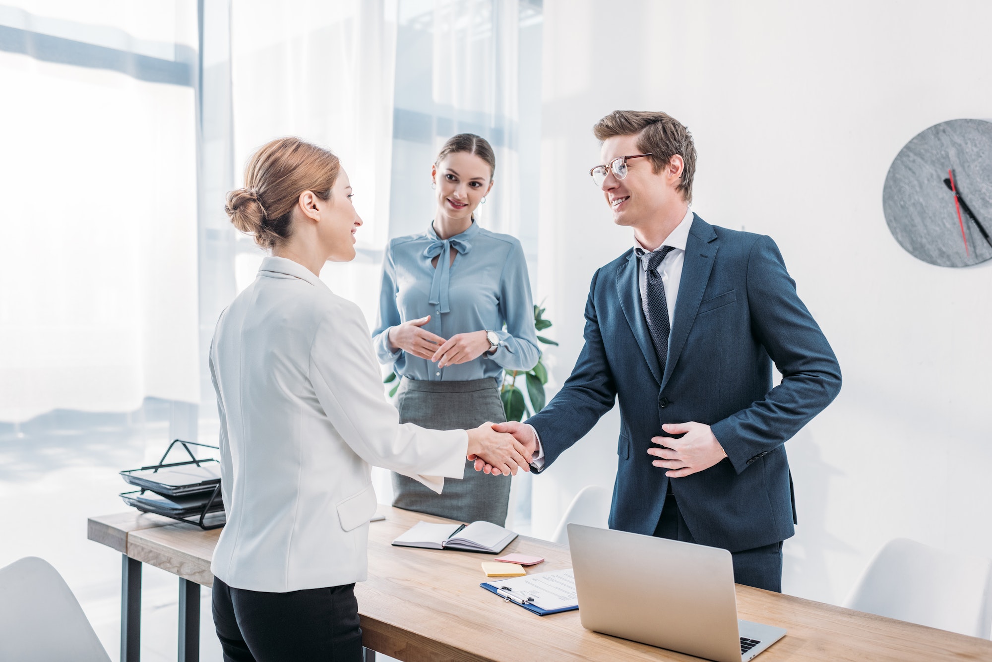 cheerful-recruiter-shaking-hands-with-woman-near-colleague-in-office